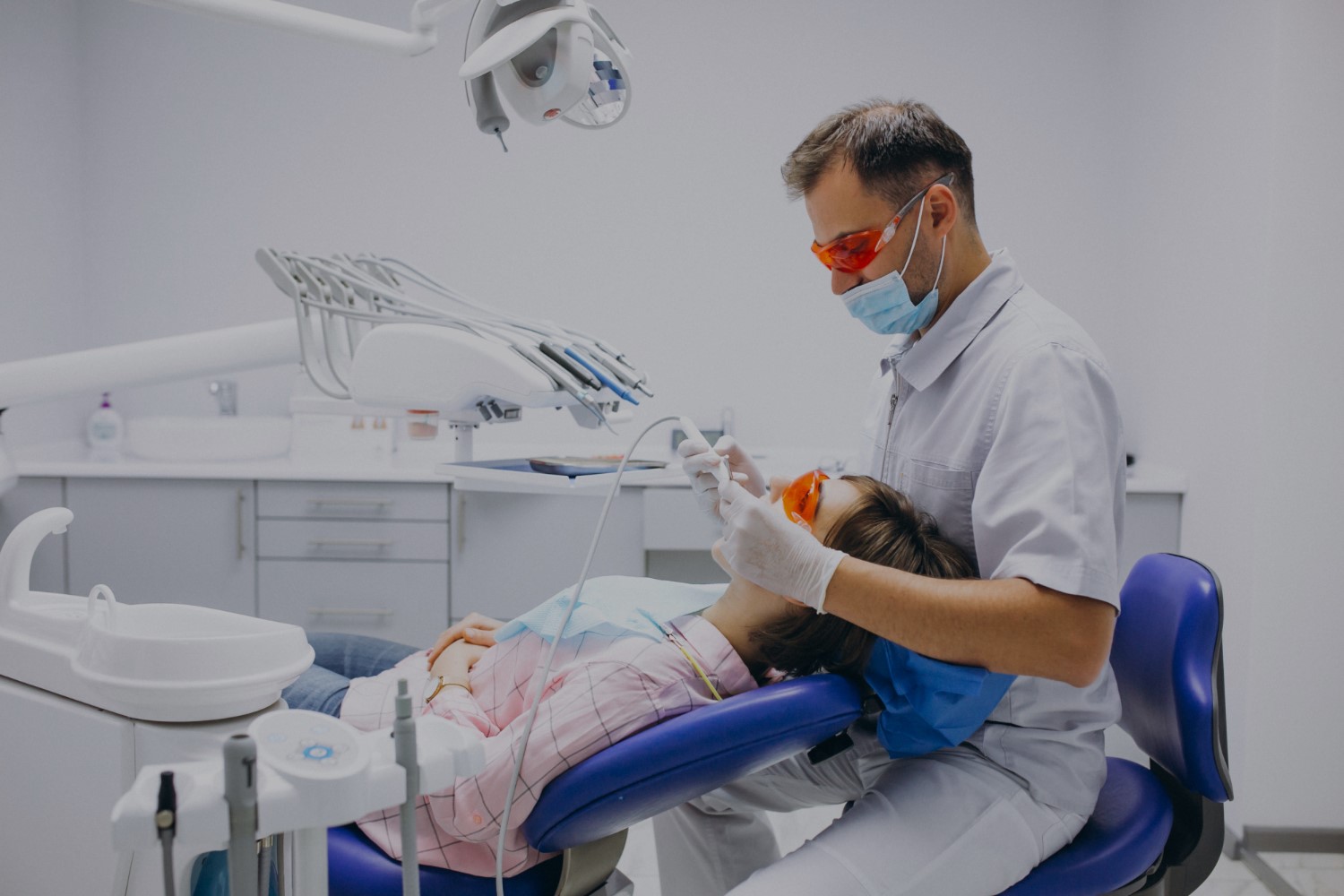 s 1 Dentist wearing protective goggles and mask performing a dental procedure on a patient reclined in a dental chair, both wearing protective eyewear.